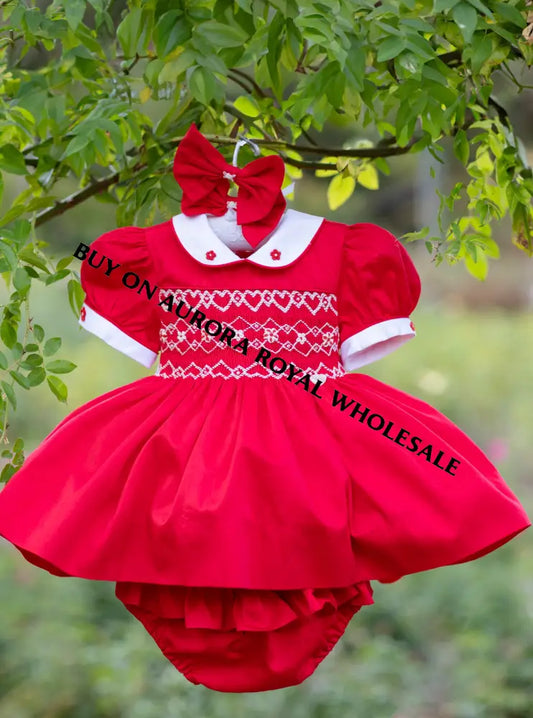 A baby girl's red dress with white collar and white smocked chest detailing, hanging from a green tree branch with a matching red bow headband. The dress features puffed short sleeves, a full ruffled skirt, and matching red ruffled bloomers.